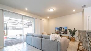 A modern living room with a grey sofa, a dining area with a wicker chair, and a large window providing natural light. - Luxury 5BR Villa - Private Pool - Water Park Resort - Near Disney (Davenport)