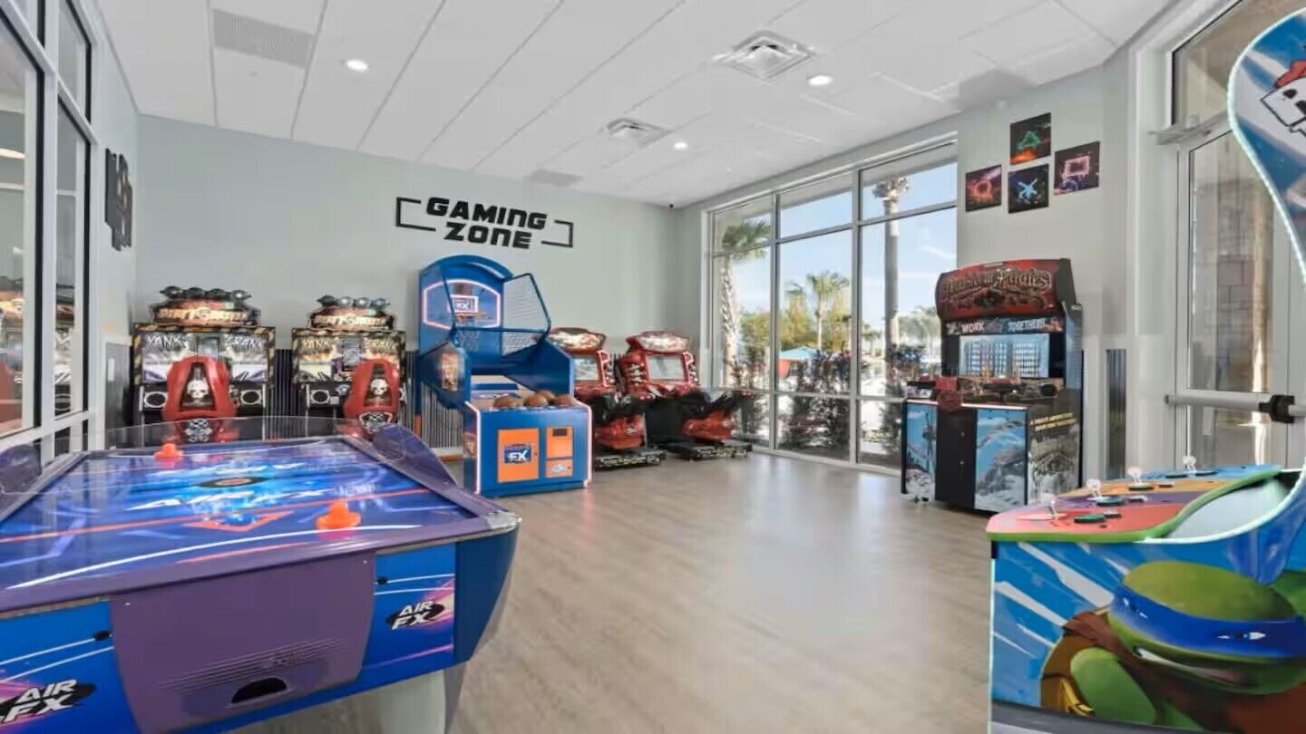 An indoor gaming area with various arcade machines, a basketball hoop, and a large window providing a view of the outside.