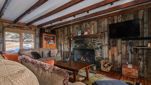 Un intérieur rustique avec une cheminée en pierre, des poutres en bois, un téléviseur et un coin salon confortable.