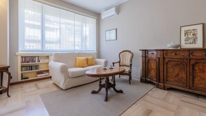Ein Wohnzimmer mit einer Couch, einem Holztisch, einem Bücherregal, einer Kommode und einem großen Fenster mit Jalousien.