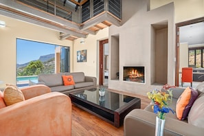 A modern living room with a fireplace, comfortable seating, and a large window overlooking a scenic view. - Houtbay Hilltop Villa (Cape Town)
