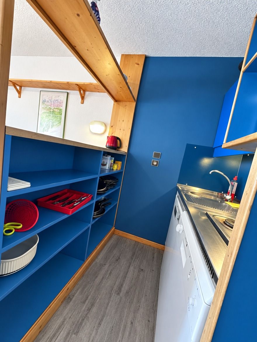 A compact kitchen with blue walls, wooden shelves, and a built-in dishwasher.