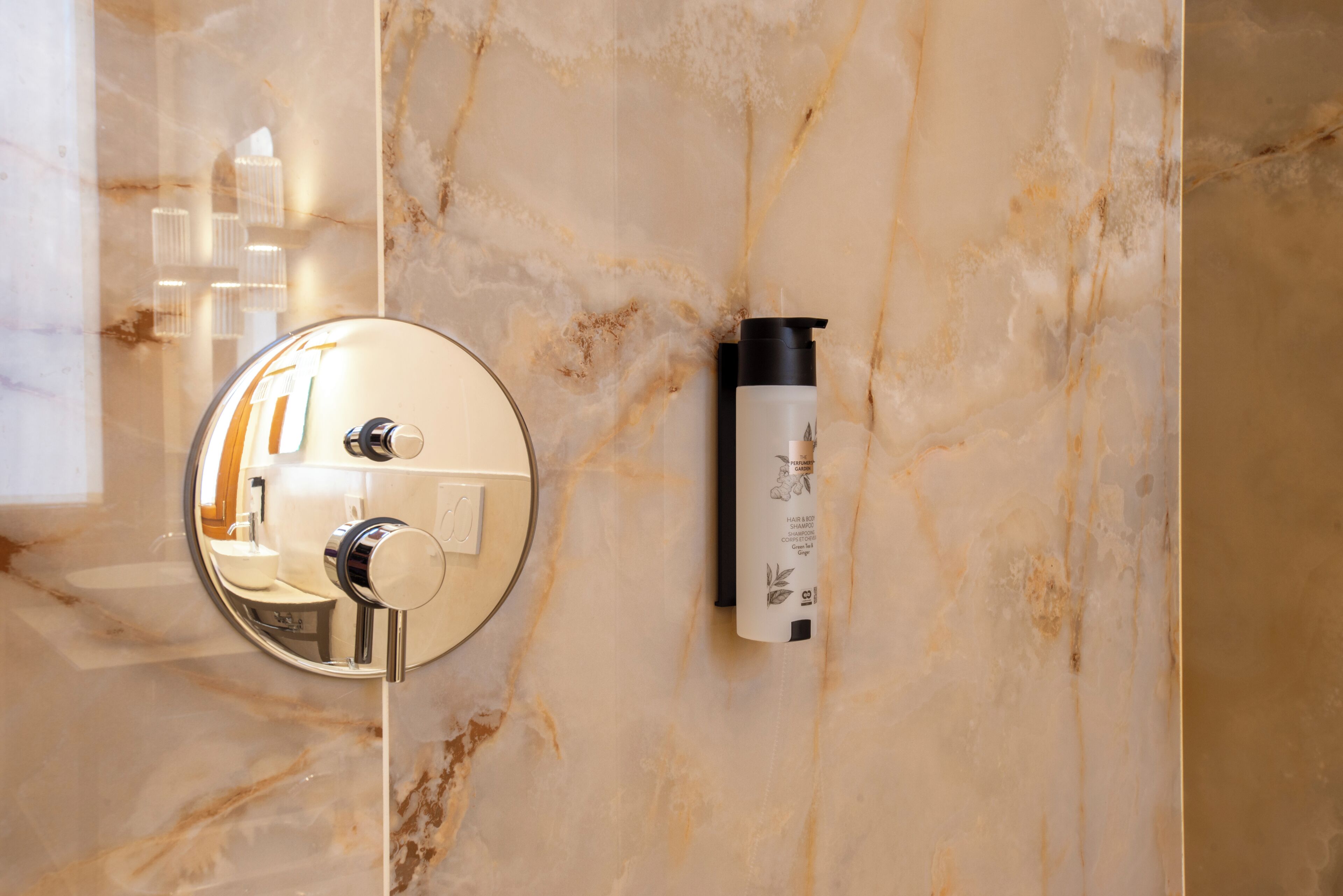 A bathroom with a marble wall, a round mirror, a faucet, and a bottle of shampoo.