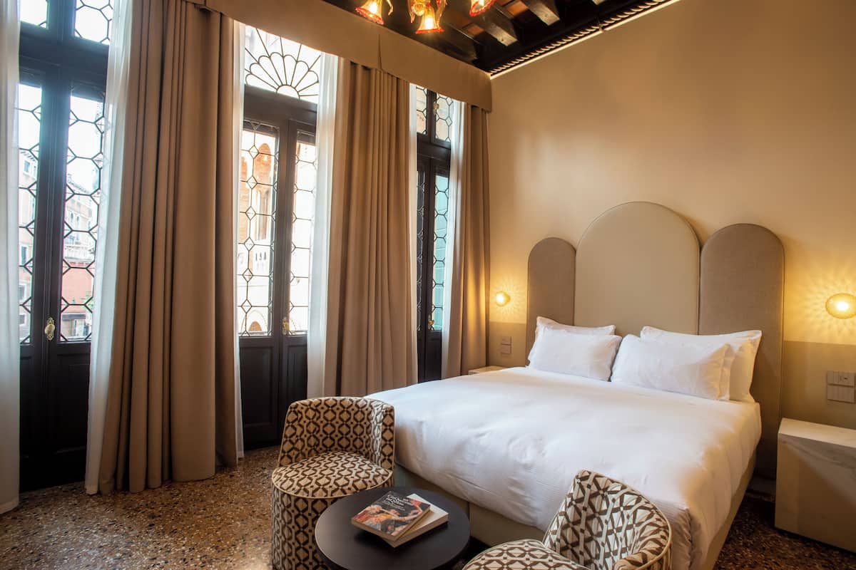 A hotel room with a large bed, two patterned armchairs, a small table with a book, and a window with curtains.