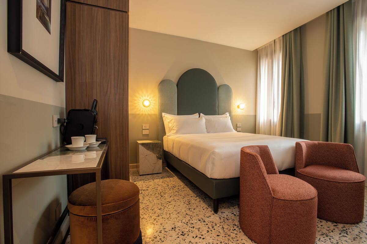 A hotel room with a bed, two chairs, a small table with cups, a trash bin, and a nightstand with a lamp.