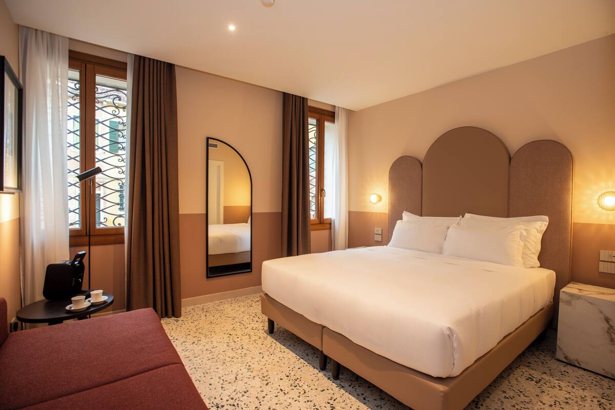 A hotel room with a large bed, a bedside table, a chair, a mirror, and a window with curtains.