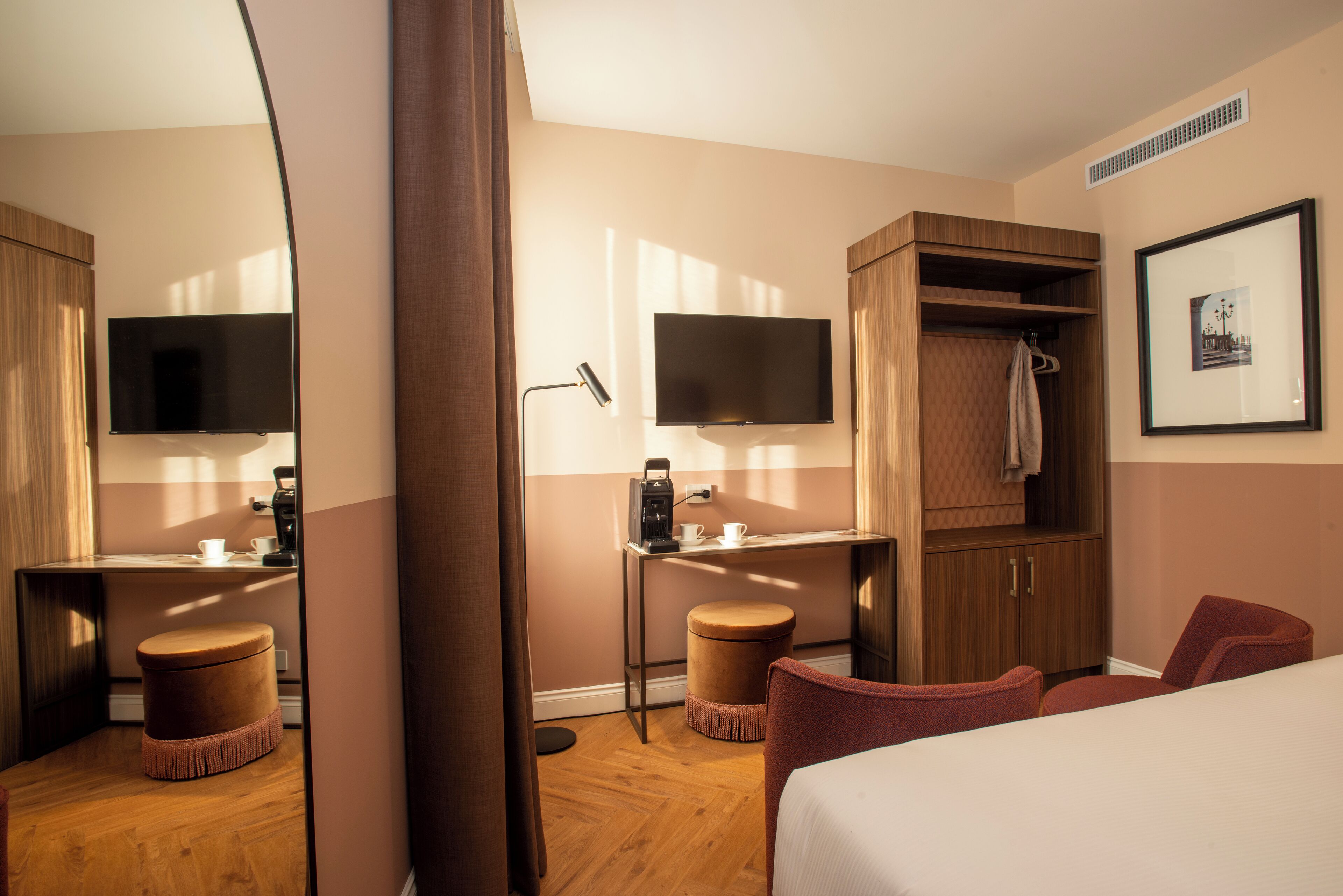A hotel room with a bed, two chairs, a desk with a TV, a coffee machine, and a wardrobe.