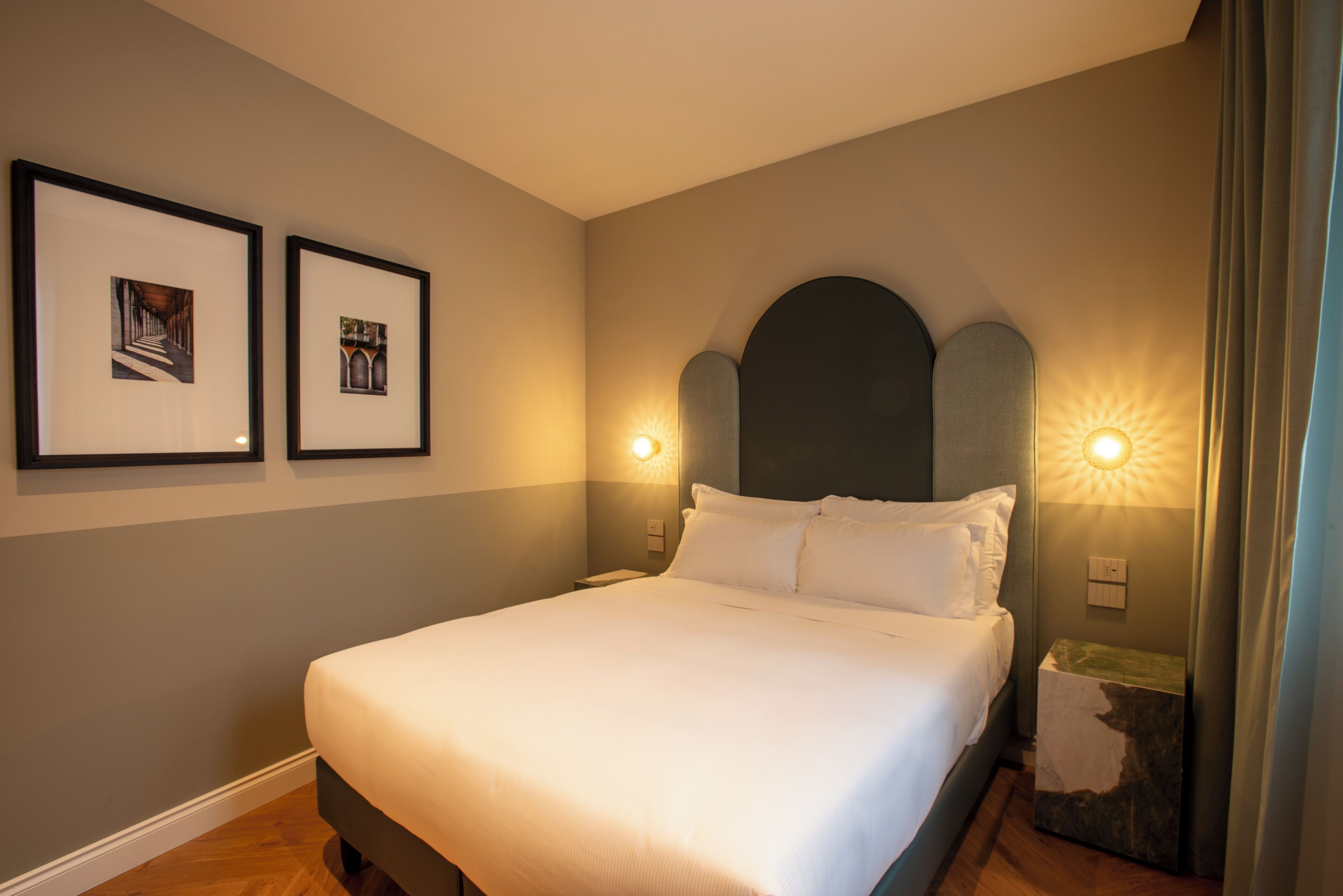 Un lit bien fait dans une chambre où l’on aperçoit deux tableaux encadrés au mur, une tête de lit avec une applique lumineuse et une table de chevet avec une lampe.