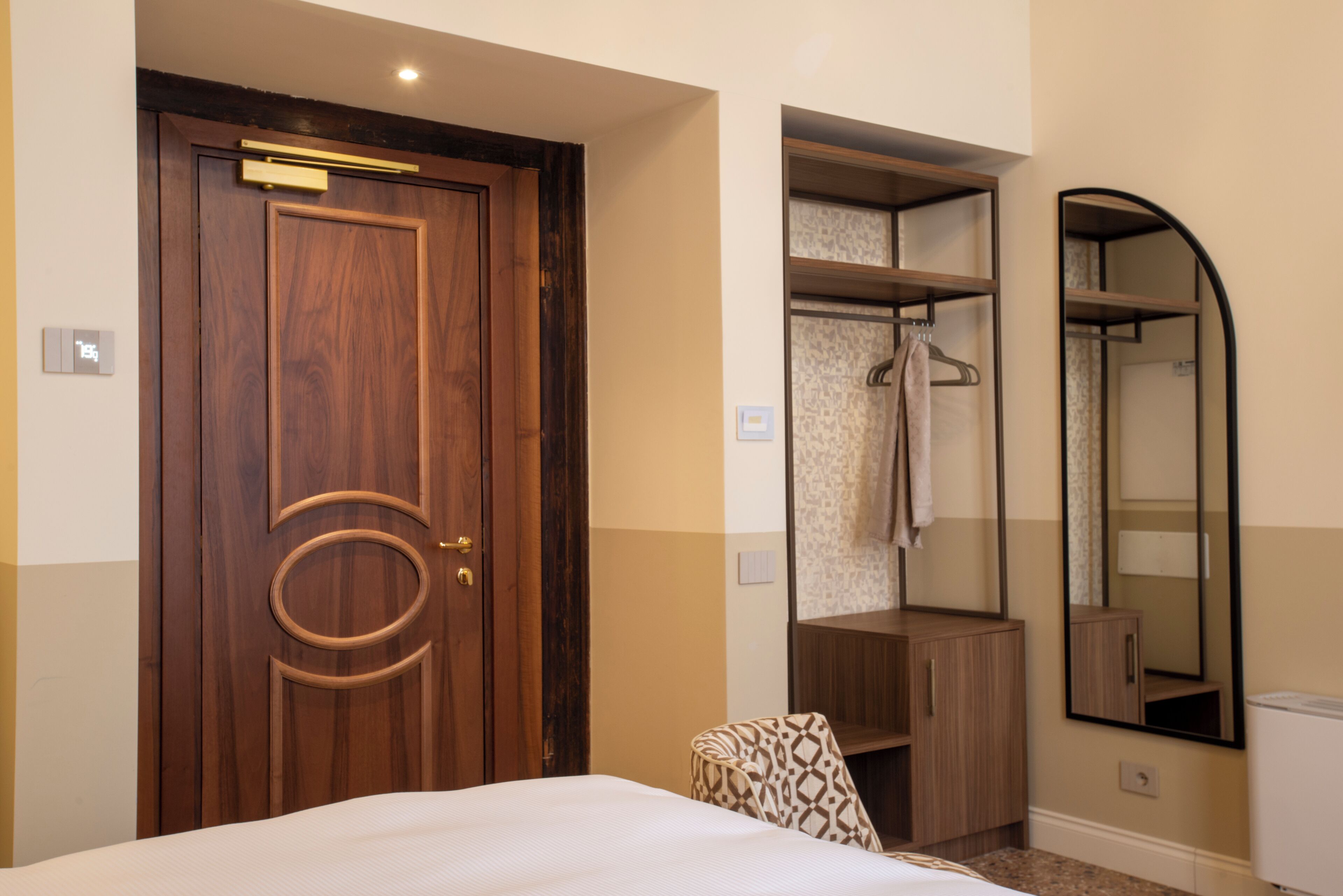 A hotel room with a wooden door, a bed, a wardrobe, a mirror, and a towel rack.