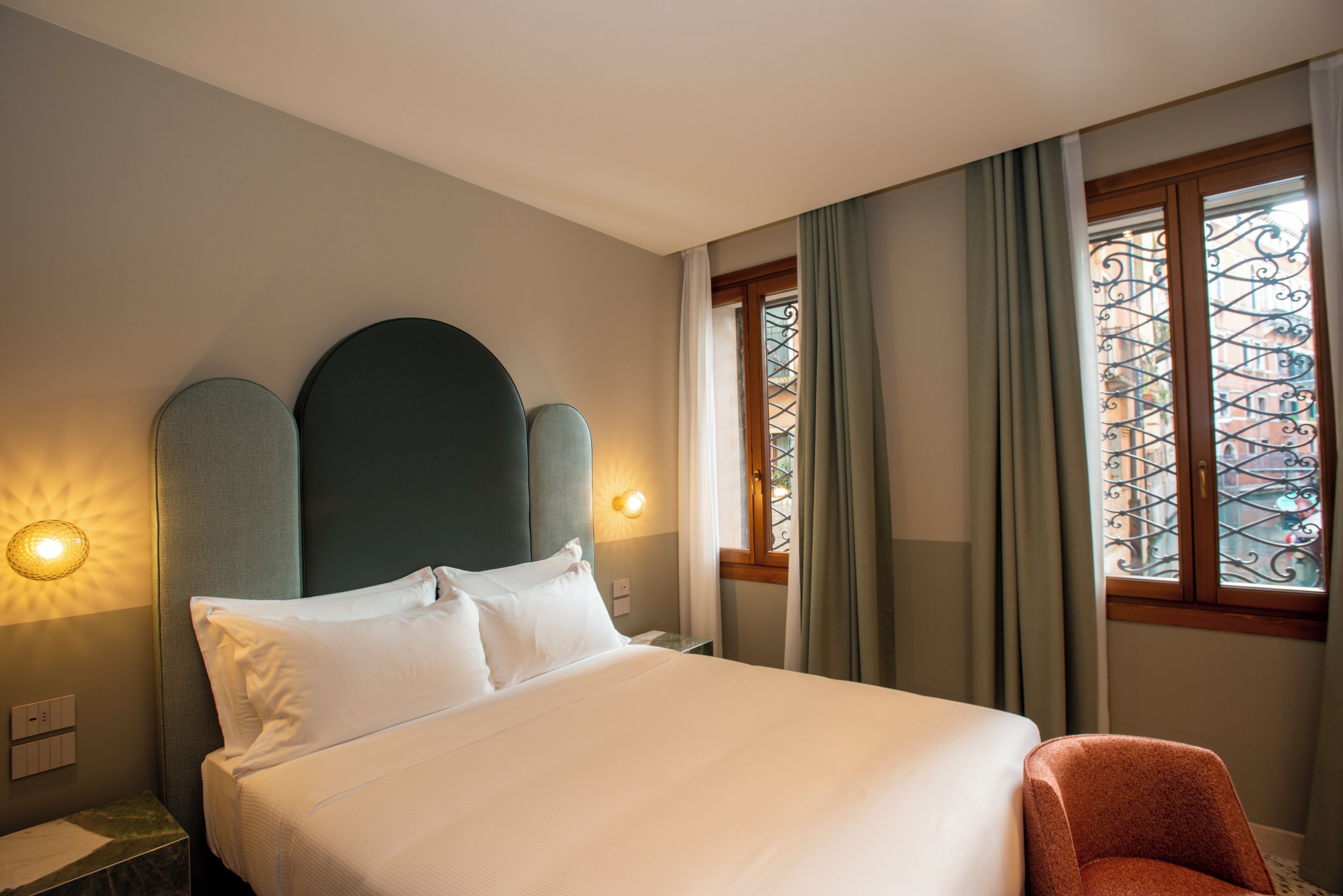 A hotel room with a bed, two bedside lamps, a nightstand, a chair, and a window with a view of buildings.