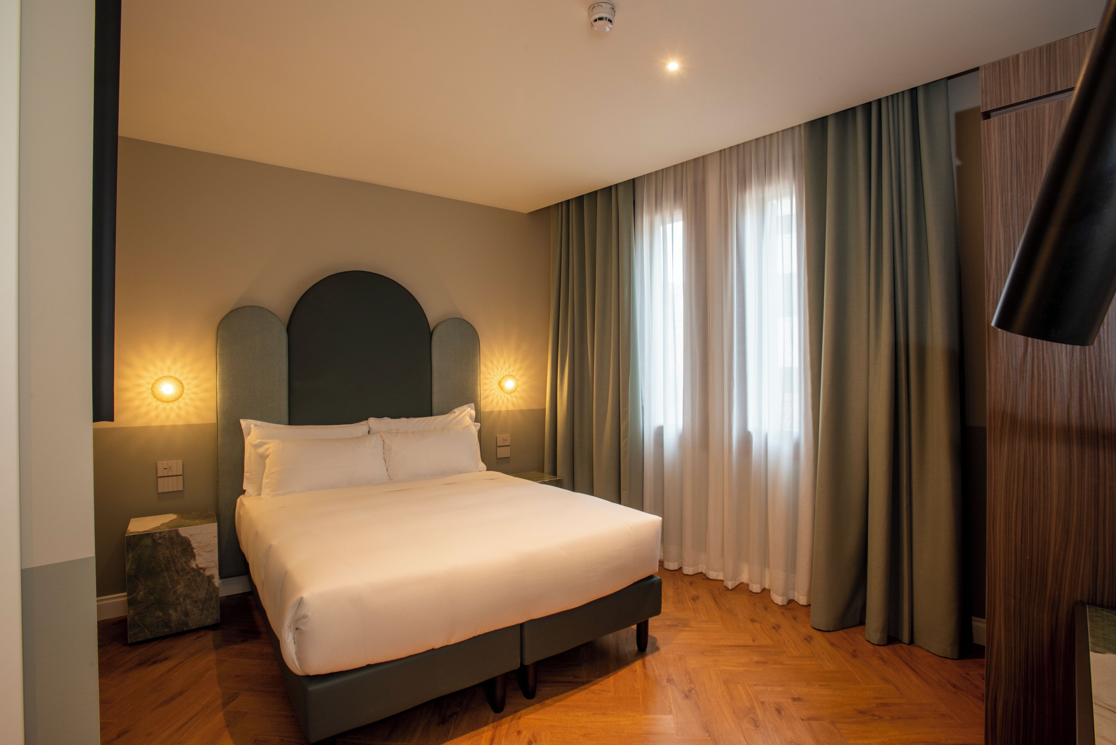 Ein modernes Hotelzimmer mit einem großen Bett, zwei Nachttischlampen, einem Flachbildfernseher und Blick ins Freie durch transparente Vorhänge.