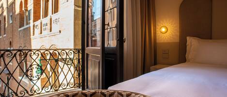 Una habitación de hotel con cama, ventana con vista a un edificio, una silla con estampado y una lámpara de pared.