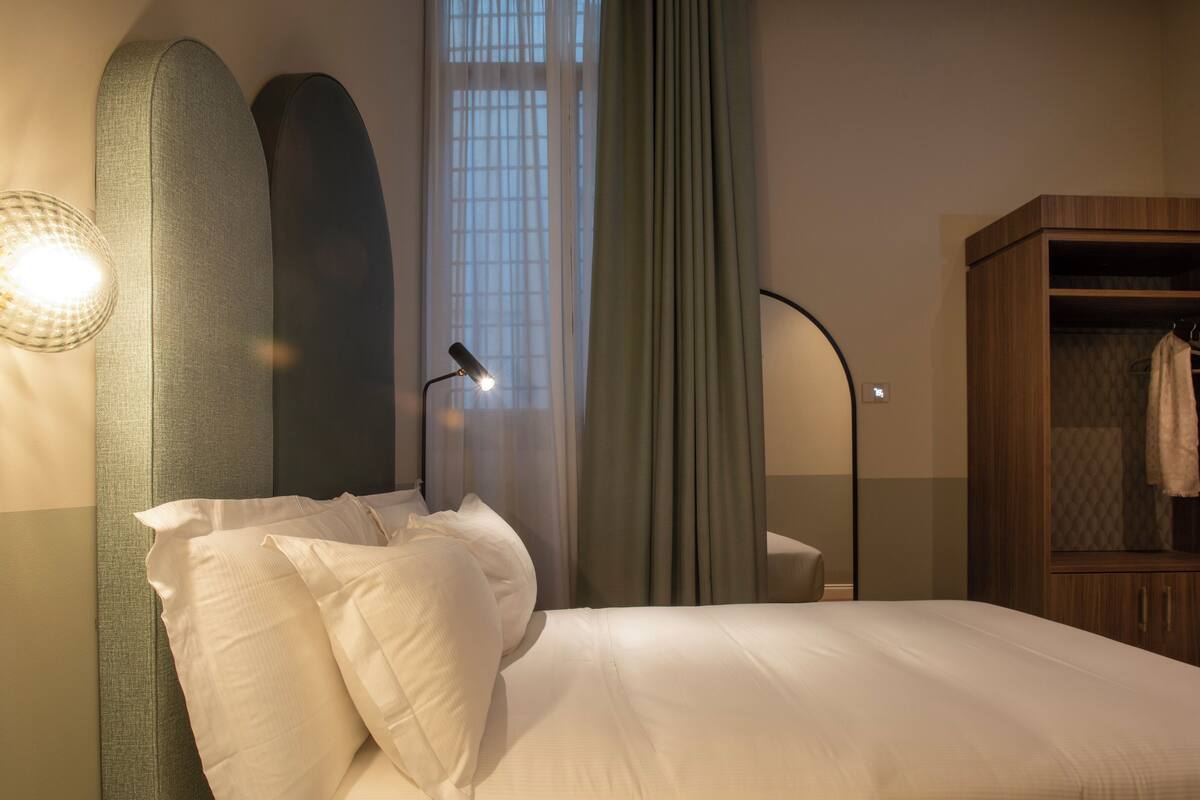 A neatly made bed with pillows, a bedside lamp, a wardrobe, and a window with curtains.