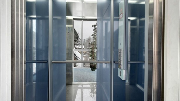 Um elevador com paredes metálicas e porta de vidro, que dá acesso a um corredor com janela.