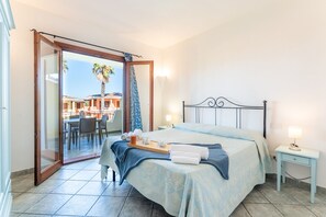 A bedroom with a bed, a bedside table, a tray with towels, and a view of an outdoor patio with tables and chairs. - Calm Escape in Seaside Resort (Palau)