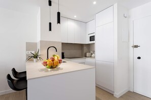 A modern kitchen with white cabinets, a central island with a fruit bowl, black pendant lights, and a microwave. - Genteel Home Pedro Teixeira (Madrid)