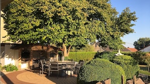 An outdoor patio with a brick floor, a table and chairs, and a large tree providing shade. - Au Jardin Du Levant" gîte with large swimming pool and games room (Holving)