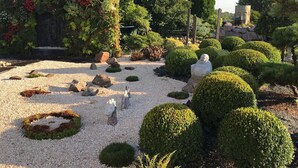 A garden with neatly trimmed bushes, a stone statue, and a variety of plants. - Au Jardin Du Levant" gîte with large swimming pool and games room (Holving)