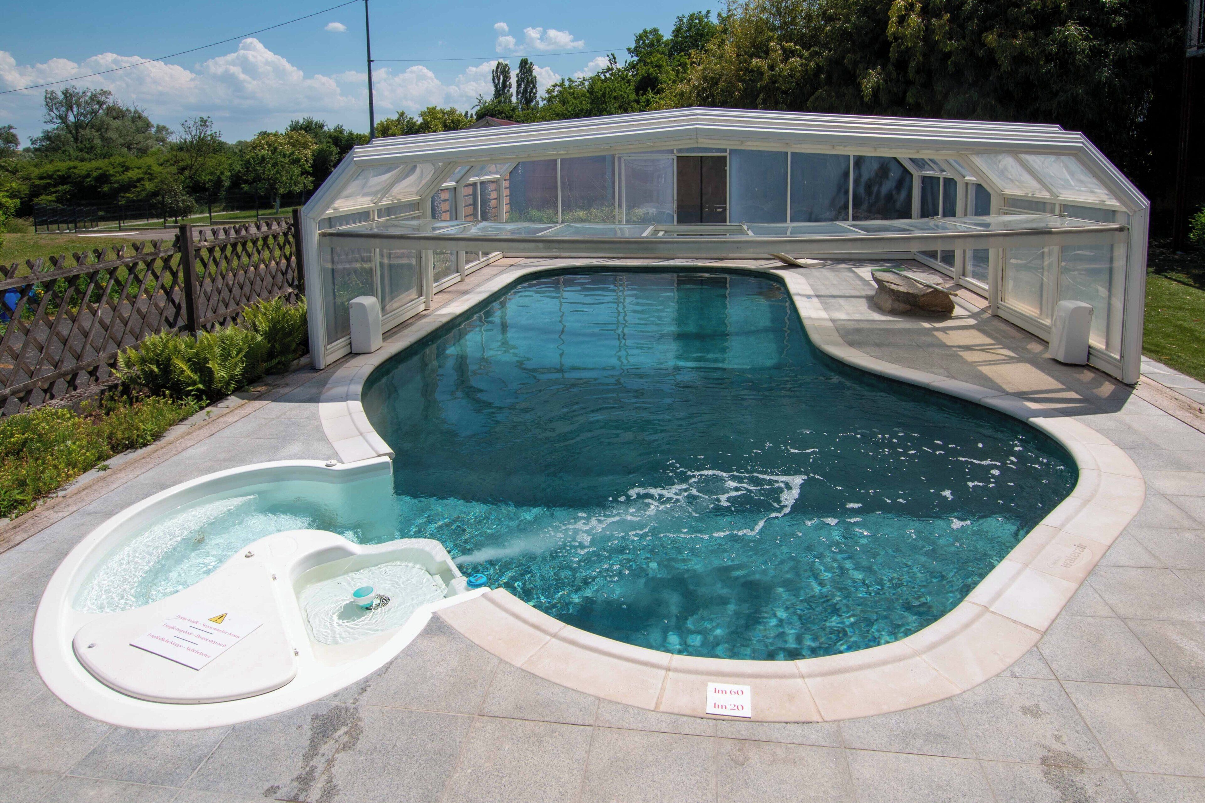A swimming pool with a glass-enclosed hot tub, surrounded by a fence and greenery.