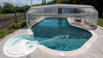A swimming pool with a glass-enclosed hot tub, surrounded by a fence and greenery.