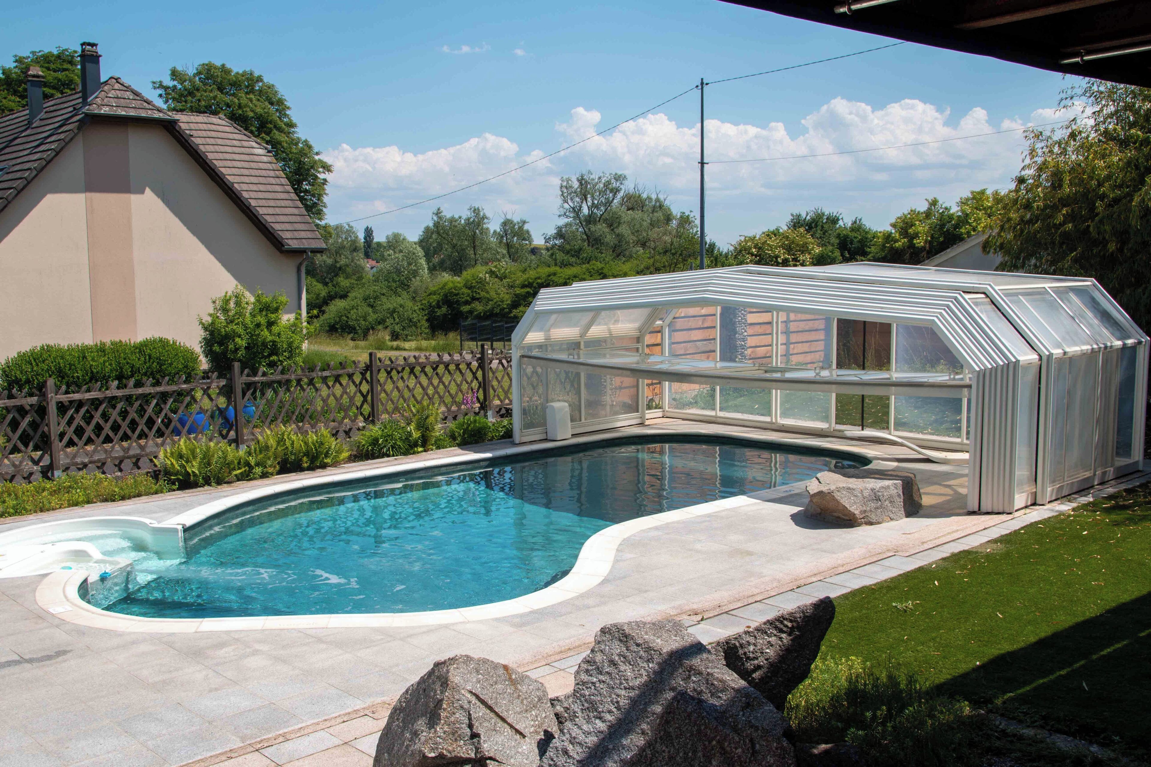 A swimming pool with a glass greenhouse structure, a house in the background, and a fenced yard.