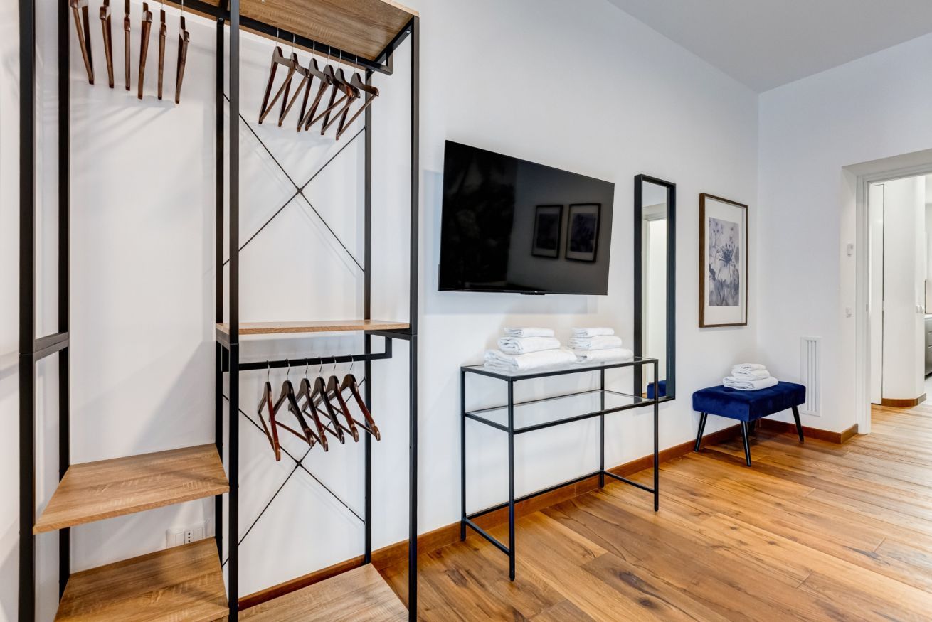 A hotel room with a wooden floor, a TV mounted on the wall, a metal rack with clothes, and a small table with towels.