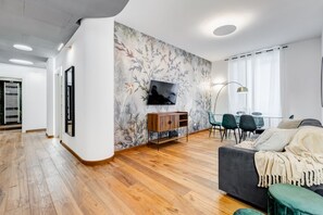 A modern living room with a wooden floor, a dark sofa, a TV mounted on the wall, and a dining area with chairs and a table. - BDC - Dragonfly 3BDR 2BATH @Testaccio (roma)