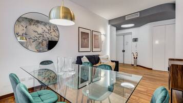 A modern living room with a glass dining table, teal chairs, a black sofa, and a large circular mirror.