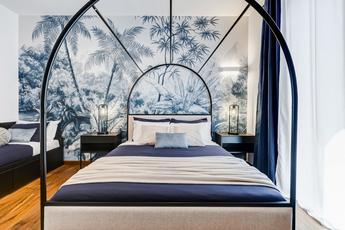 A bedroom with a canopy bed, a mural of tropical foliage, and bedside lamps.