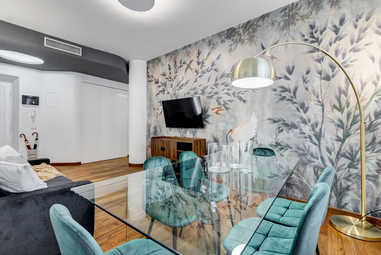 A modern living room with a glass coffee table, teal chairs, a black sofa, a wooden cabinet, a flat-screen TV, and a large floor lamp.