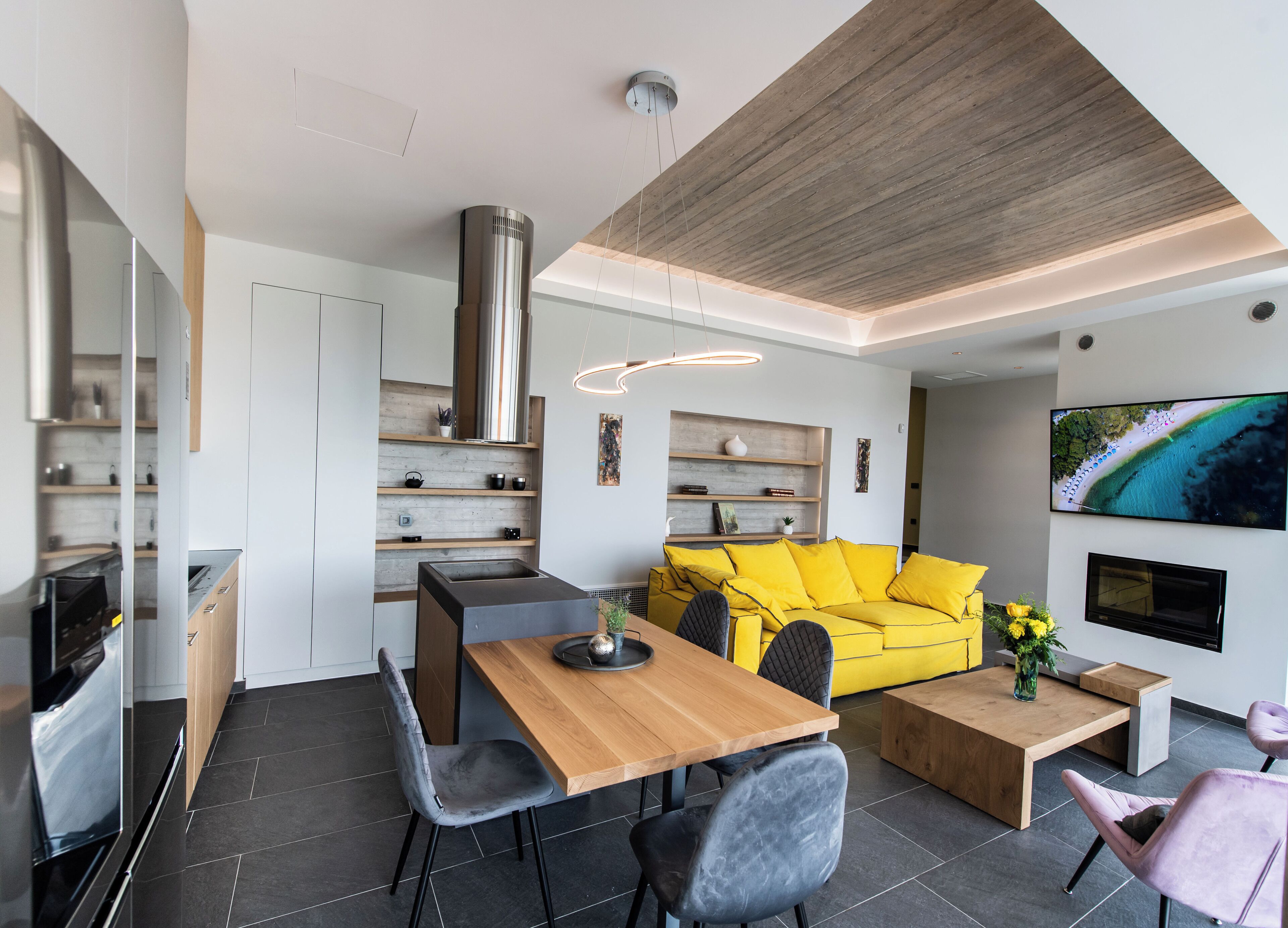 Une cuisine moderne dotée d’une cuisinière intégrée, un coin repas avec une table et des chaises en bois, un canapé jaune vif et un téléviseur à écran plat fixé au mur.
