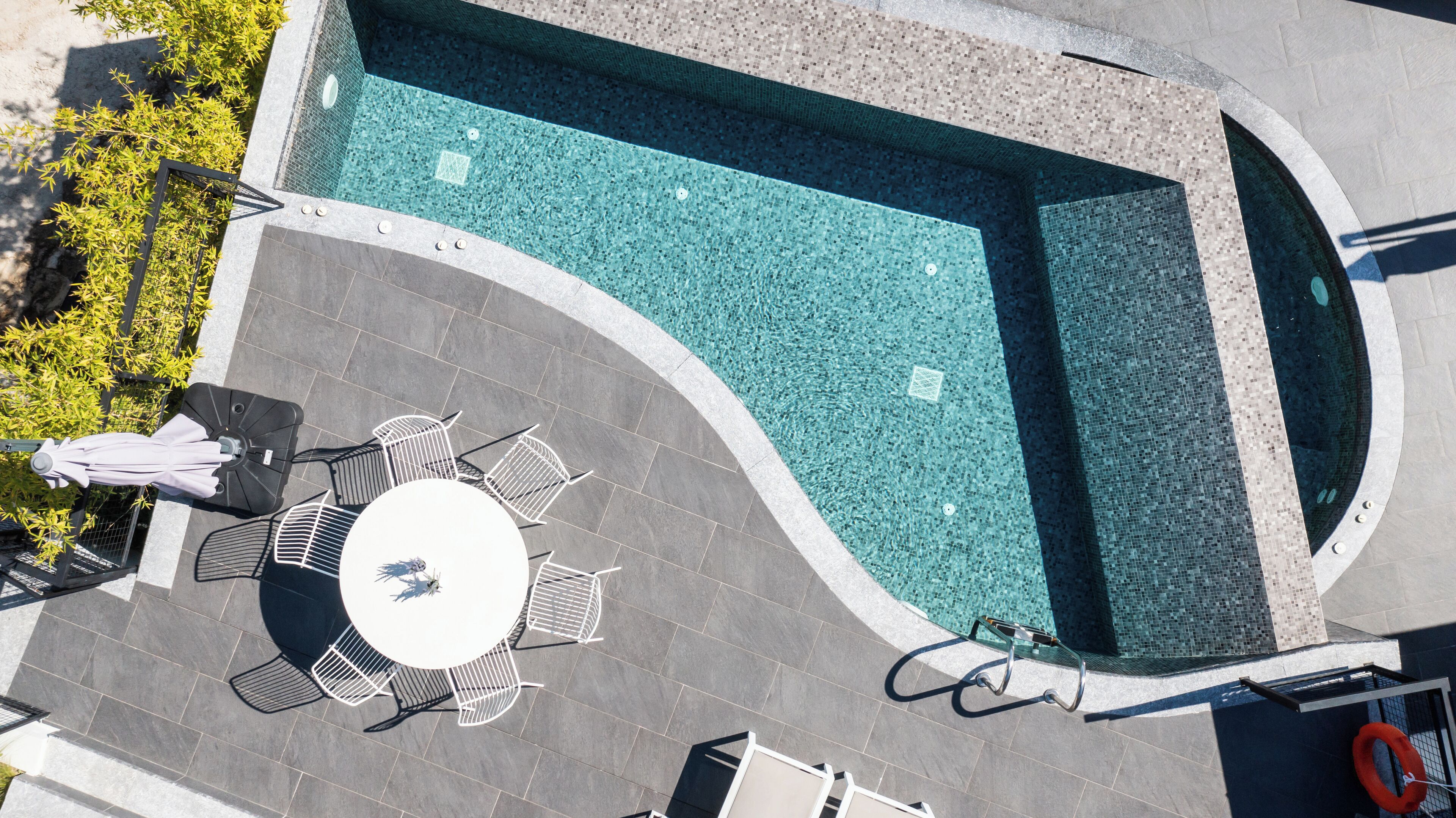 An aerial view of a swimming pool with a tiled deck, a round table with chairs, and a lifebuoy.