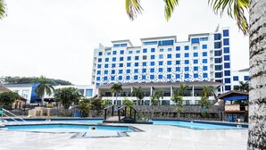 Un hotel moderno con zona de piscina, que presenta un edificio grande de fachada azul y blanca, palmeras y cielo despejado.