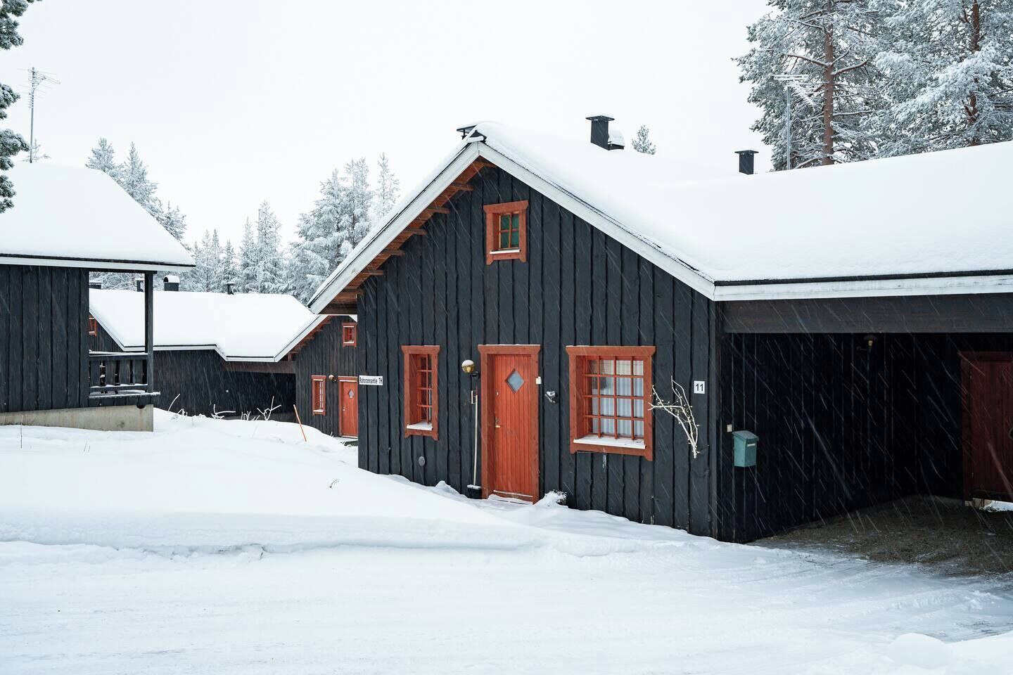 A black wooden building with a snow-covered roof, a red door, and a small window.