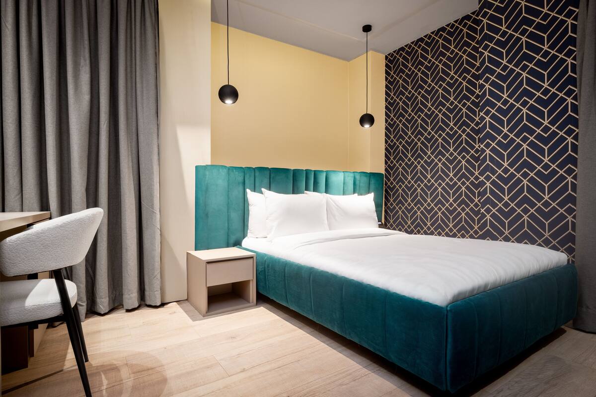 A modern hotel room with a teal bed, a small bedside table, a chair, and a patterned wall.