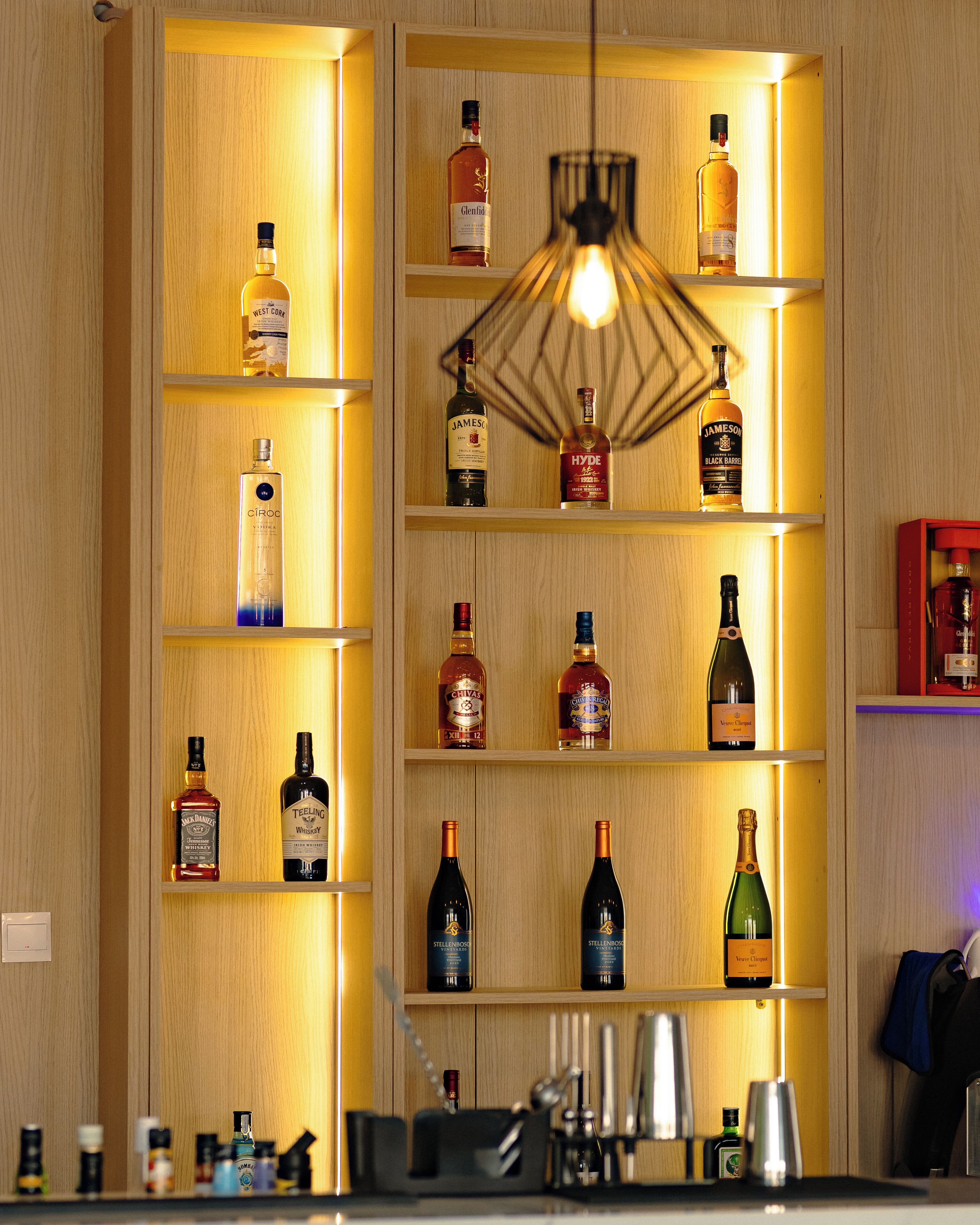 A well-lit bar area with shelves of various liquor bottles, a hanging light fixture, and bar tools on the counter.