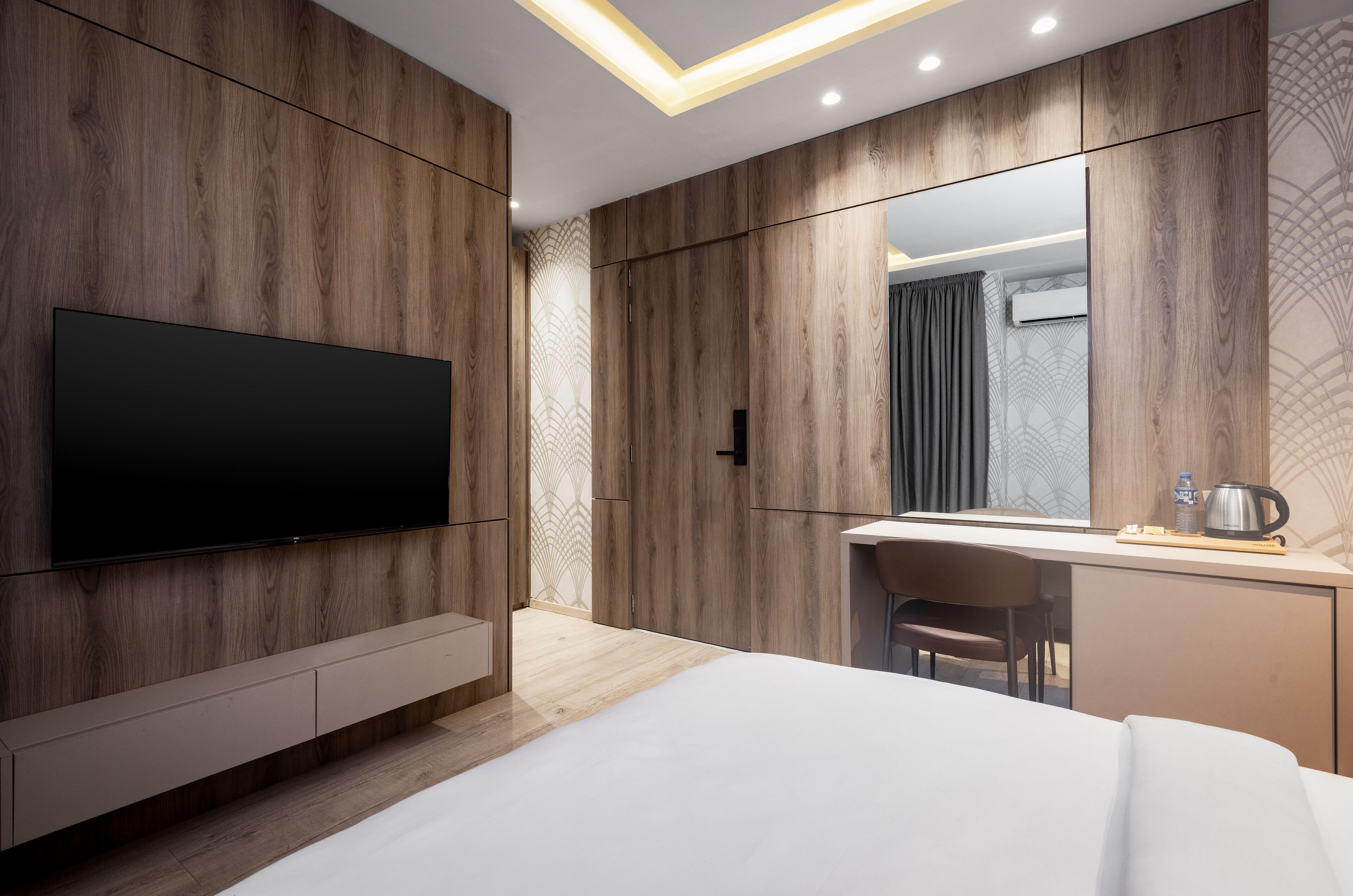 A modern hotel room with a large flat-screen TV, wooden panel walls, a desk with a chair, and a bed.