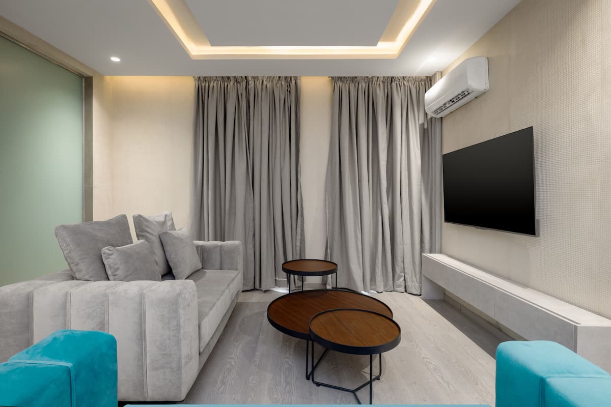 A modern living room with a grey sofa, two round wooden tables, a flat-screen TV mounted on the wall, and grey curtains.
