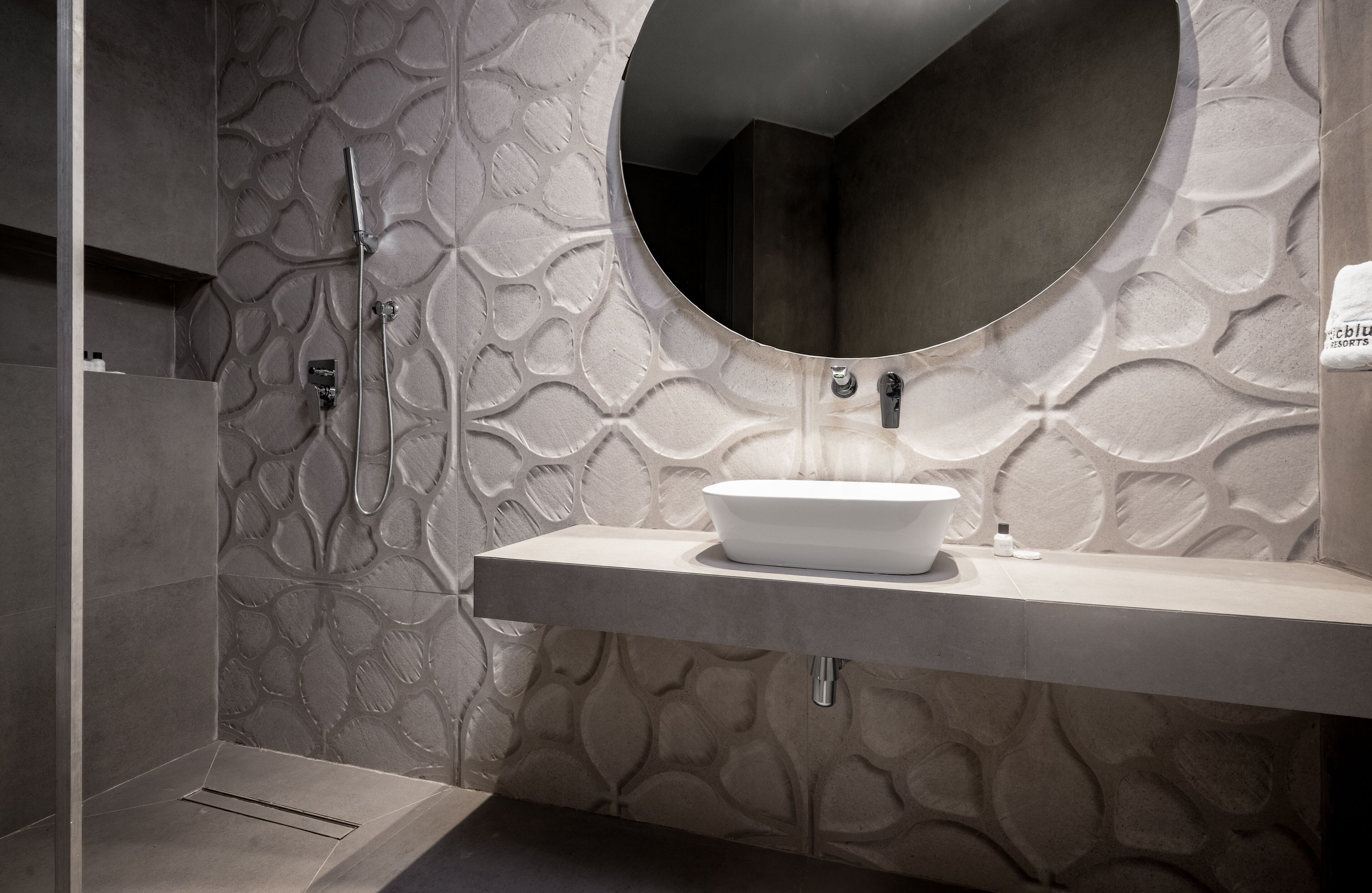 A bathroom with a round mirror, a white sink, and a shower with a textured wall.