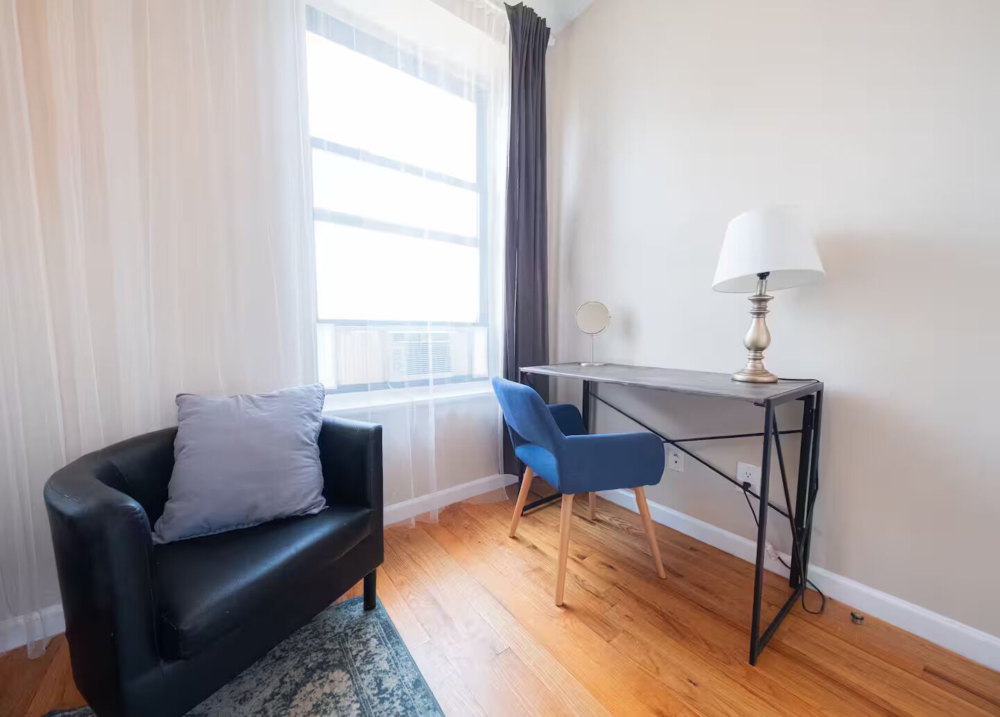Une pièce avec un canapé en cuir noir, un fauteuil bleu, un bureau avec une lampe et une fenêtre avec des rideaux.