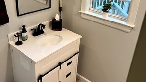 A bathroom with a white vanity, a mirror, a window with a view of trees, and a potted plant on the windowsill. - Cozy Cottage in Union, SC (Union)