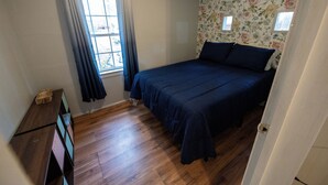 A bedroom with a wooden floor, a bed with dark bedding, a window with curtains, and floral wallpaper. - Cozy Cottage in Union, SC (Union)
