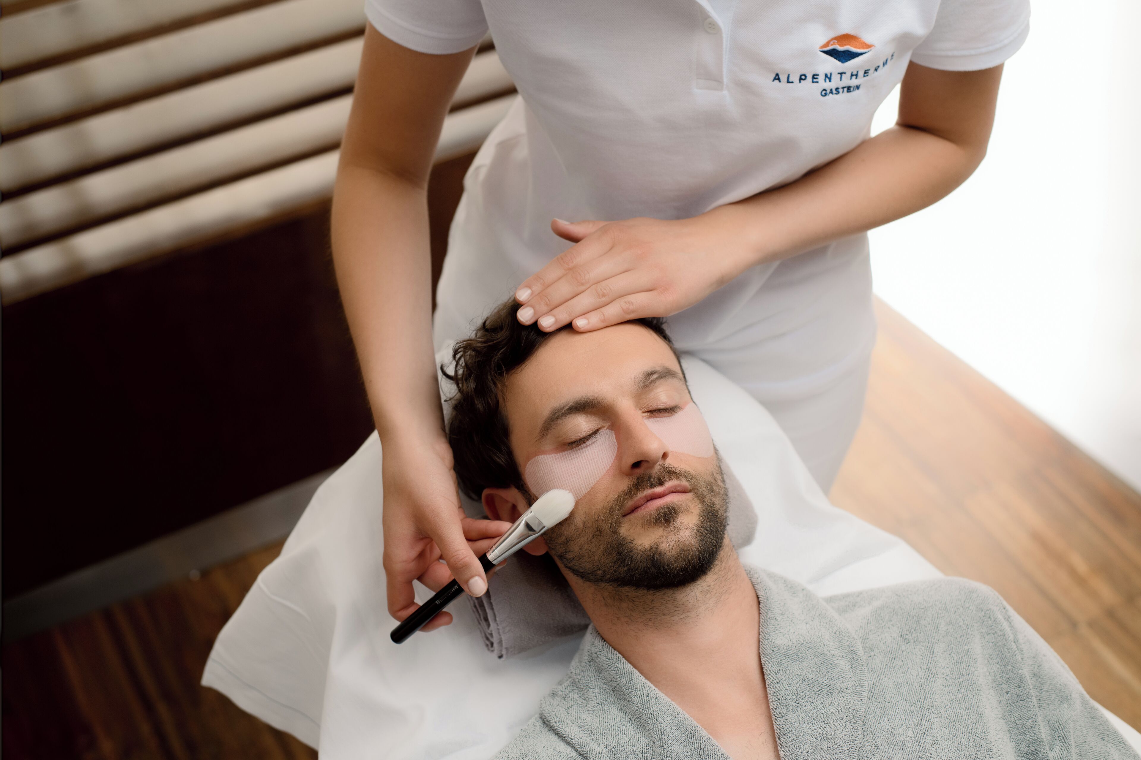A person receiving a facial treatment in a room with a wooden floor and a white wall.