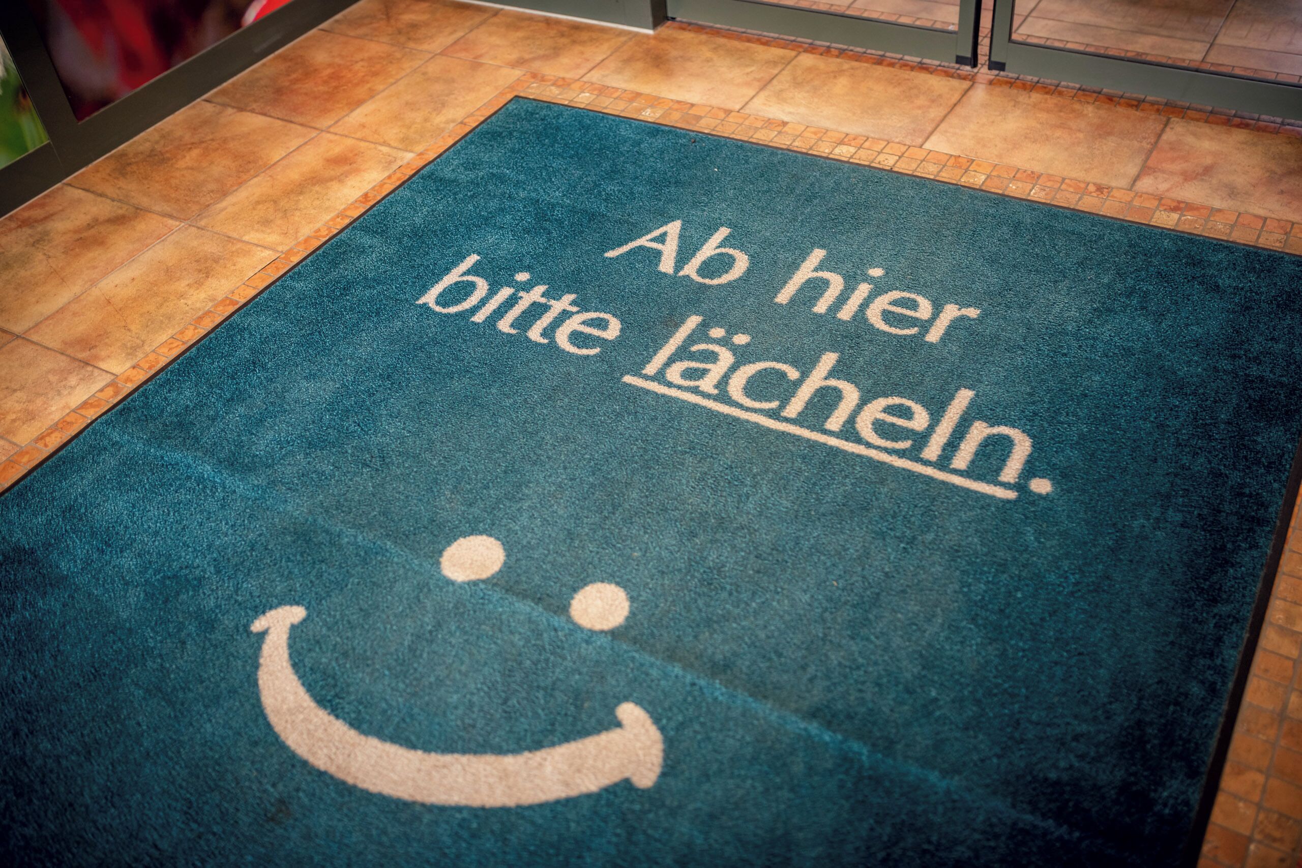A blue mat with the text "Ab hier bitte lächeln." and a smiley face.