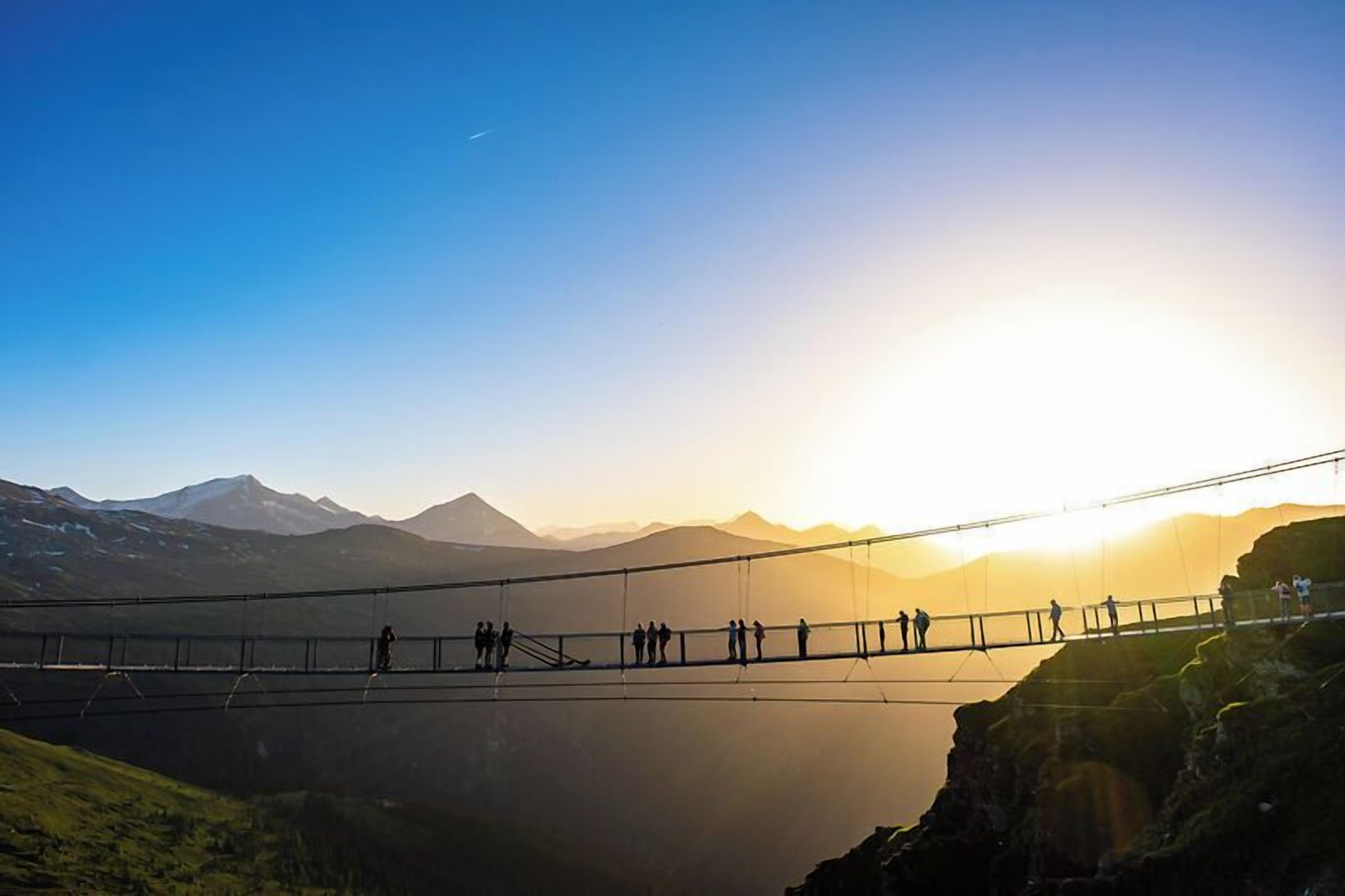 A suspension bridge with people walking across it, overlooking a valley with mountains in the background.