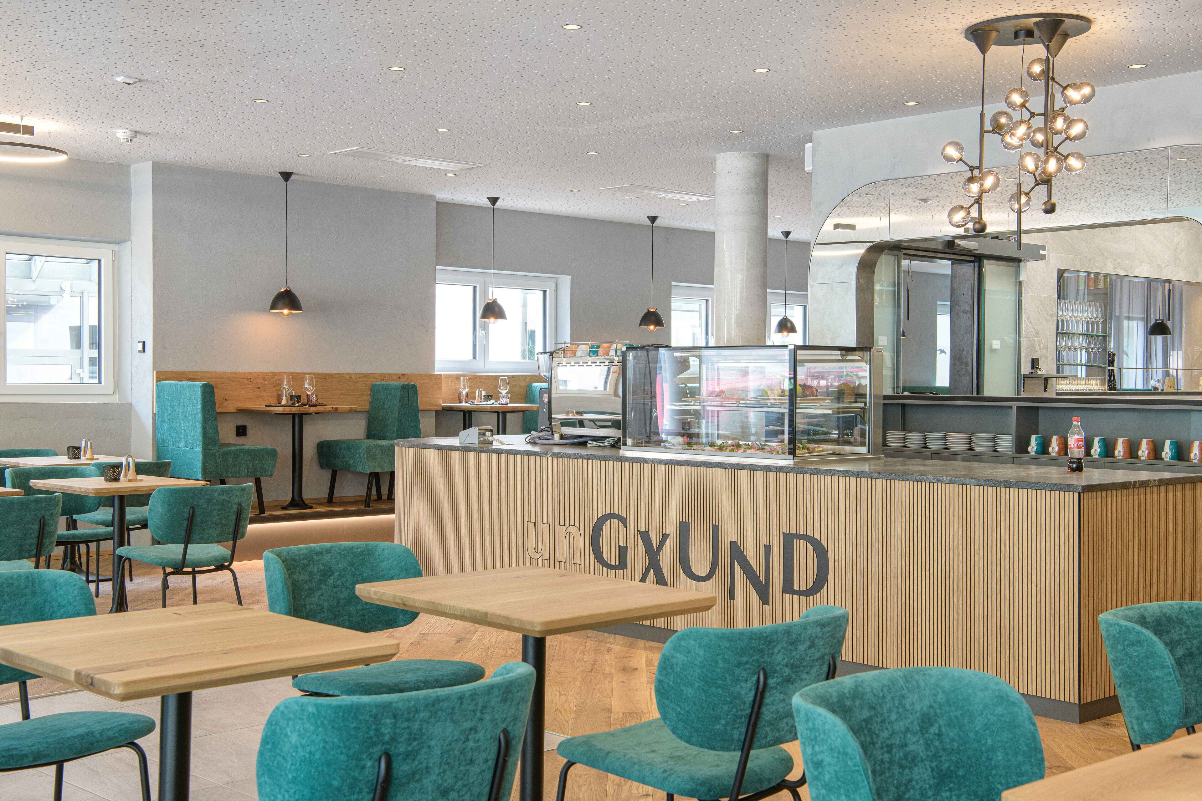 Un café moderno con sillas color turquesa y mesas de madera, una barra central con la marca 'unGxUND' y una vitrina de vidrio.
