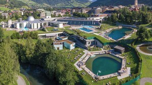 A resort with multiple swimming pools, a large building with a dome, and a church in the background. - Aktiv- & Gesundheitsresort das GXUND (Bad Hofgastein)