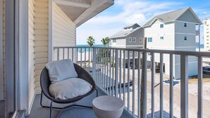 A balcony with a wicker chair and a round table, overlooking a beach and neighboring buildings. - El Mar Townhome - Unit D (Indian Shores)