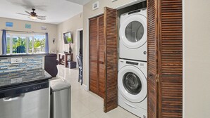 Une buanderie équipée de deux machines à laver superposées, d’un ventilateur de plafond et offrant une vue sur l’extérieur à travers une fenêtre agrémentée de rideaux.