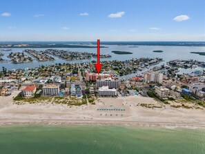 Une vue aérienne d’une ville côtière, où un hôtel imposant est mis en évidence par une flèche rouge, et qui est entouré de zones résidentielles et de plans d’eau.